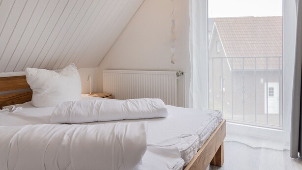 A bedroom with a sloped ceiling, a wooden bed frame, white bedding, a bedside table with a lamp, and a window with a view of a building outside.