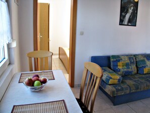 Un coin repas avec une table, des chaises et un saladier de fruits. On aperçoit un canapé avec des coussins à motifs à l’arrière-plan.