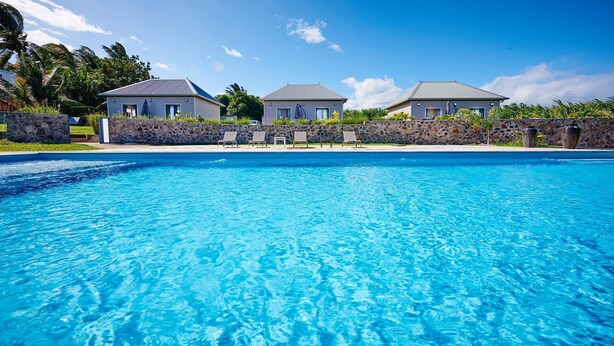 Une piscine à l’eau d’un bleu limpide, entourée d’un mur de pierre et bordée de trois maisons aux toits gris.