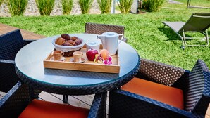 Une table ronde en verre, sur laquelle se trouve un plateau de nourriture comprenant du pain, des fruits et une théière, est placée à l’extérieur, entourée de chaises en osier, et offre une vue sur une piscine et un espace verdoyant.