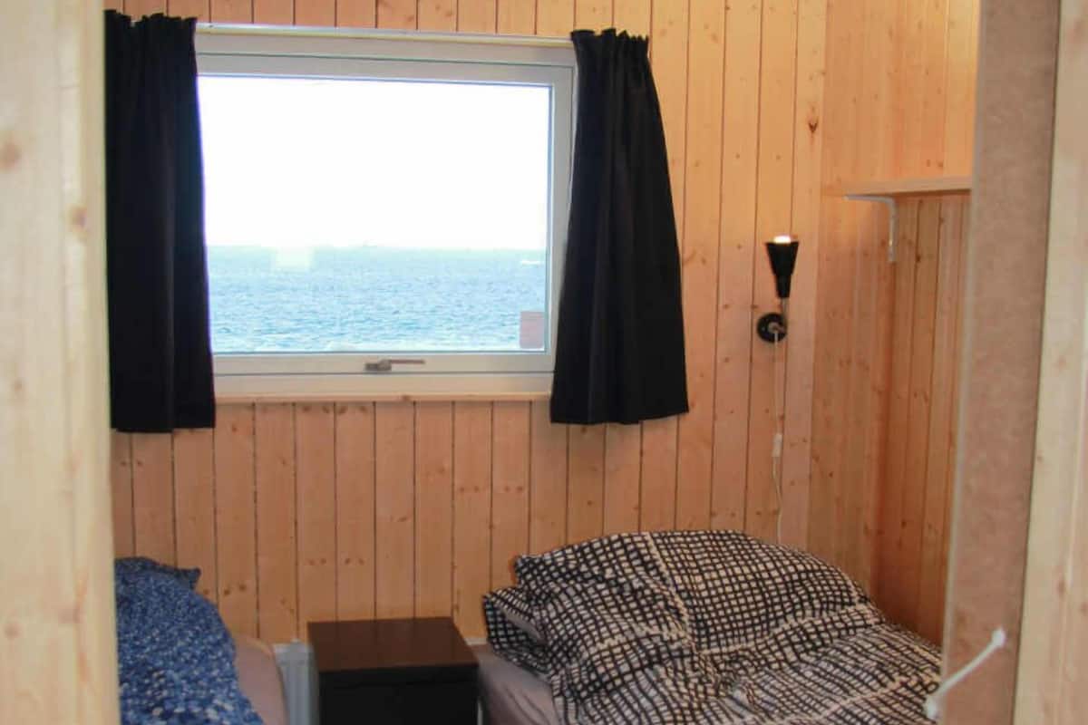 A small cabin room with wooden walls, a window with curtains, a bed with patterned bedding, and a small table.