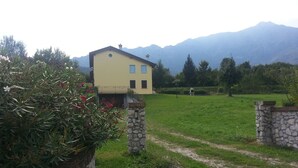 A yellow house with a stone wall entrance, a grassy field, and mountains in the background. - The house in Old Railway 2 (the places of the Great War) (Velo D'astico)