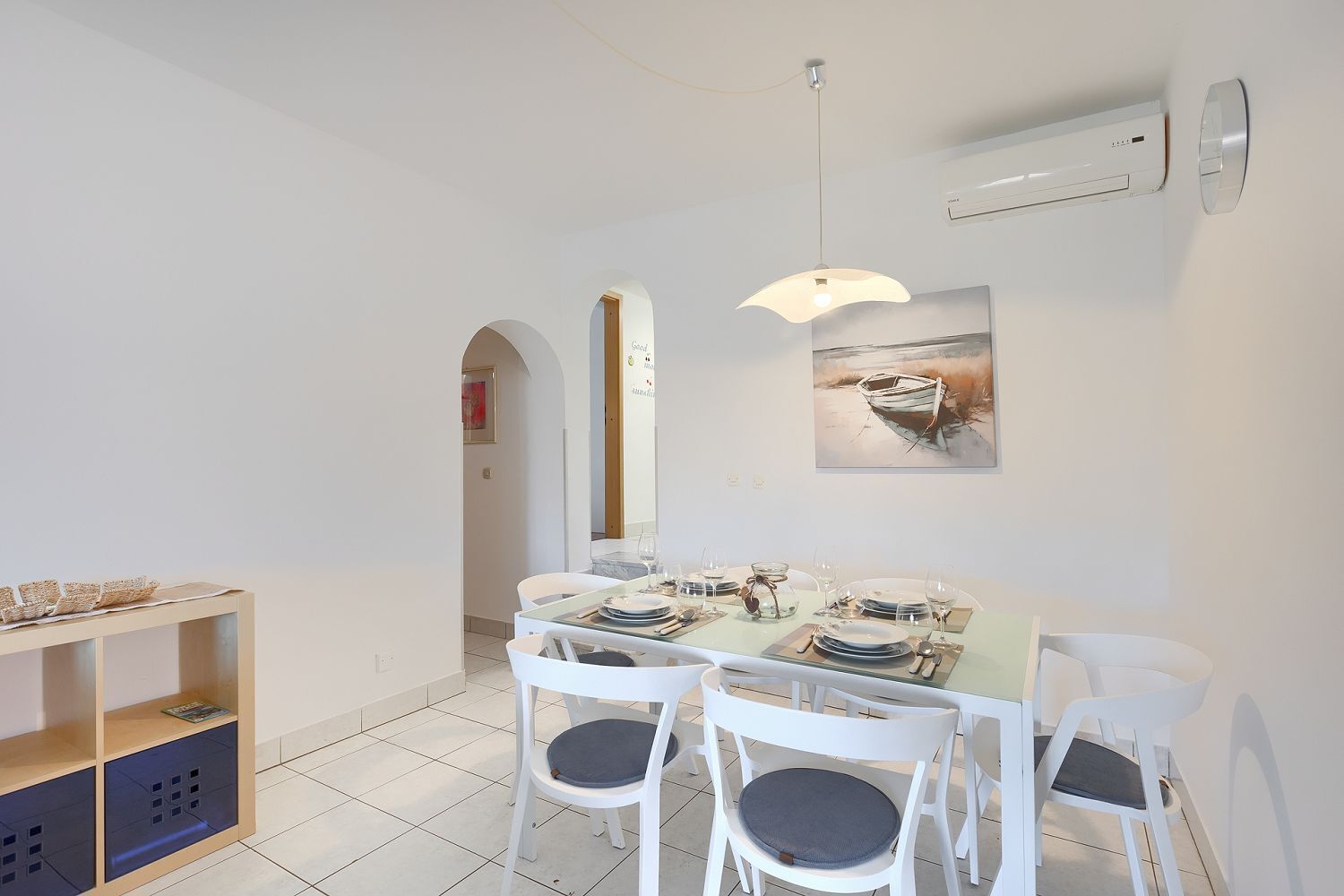 Een eetruimte met een tafel voor vier personen, witte stoelen, een hanglamp en een schilderij aan de muur.