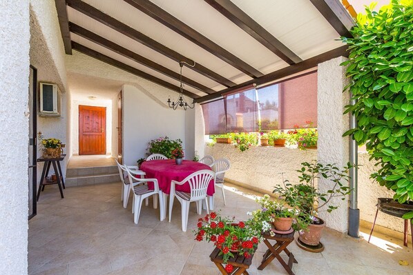Um alpendre coberto com mesa e cadeiras, plantas em vasos e vista para a rua.