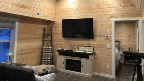 A cozy room with wooden walls, a flat-screen TV mounted above a fireplace, a ladder, and a window with a view. - Eagle Lake Fishing - Getaway Cabin (Waldhof)