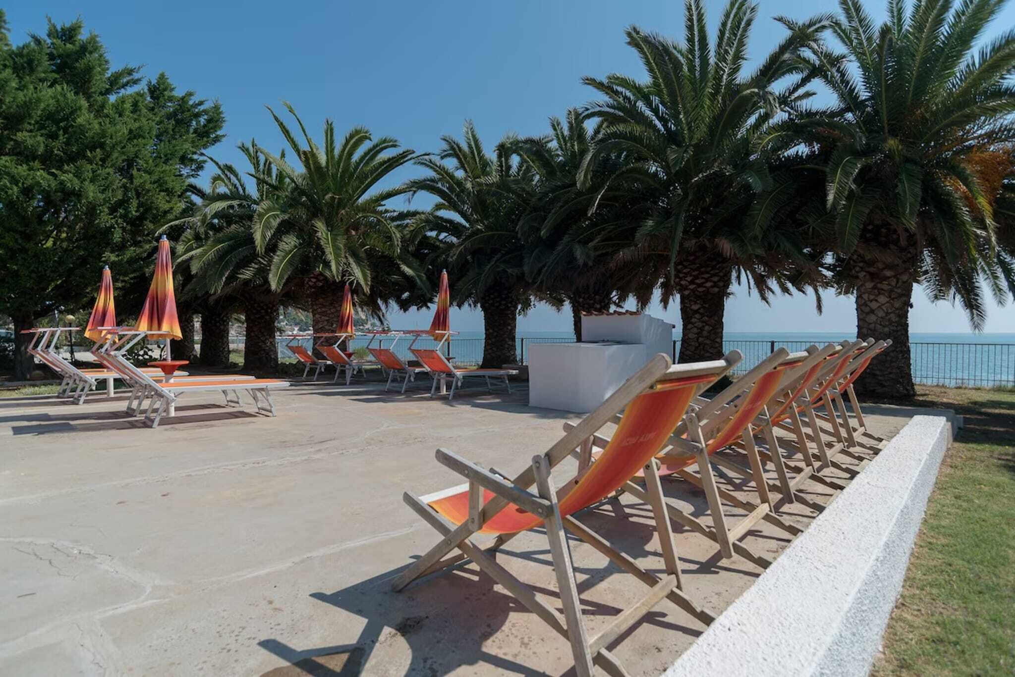 A beachside lounge area with sun loungers and umbrellas, palm trees, and a view of the sea.