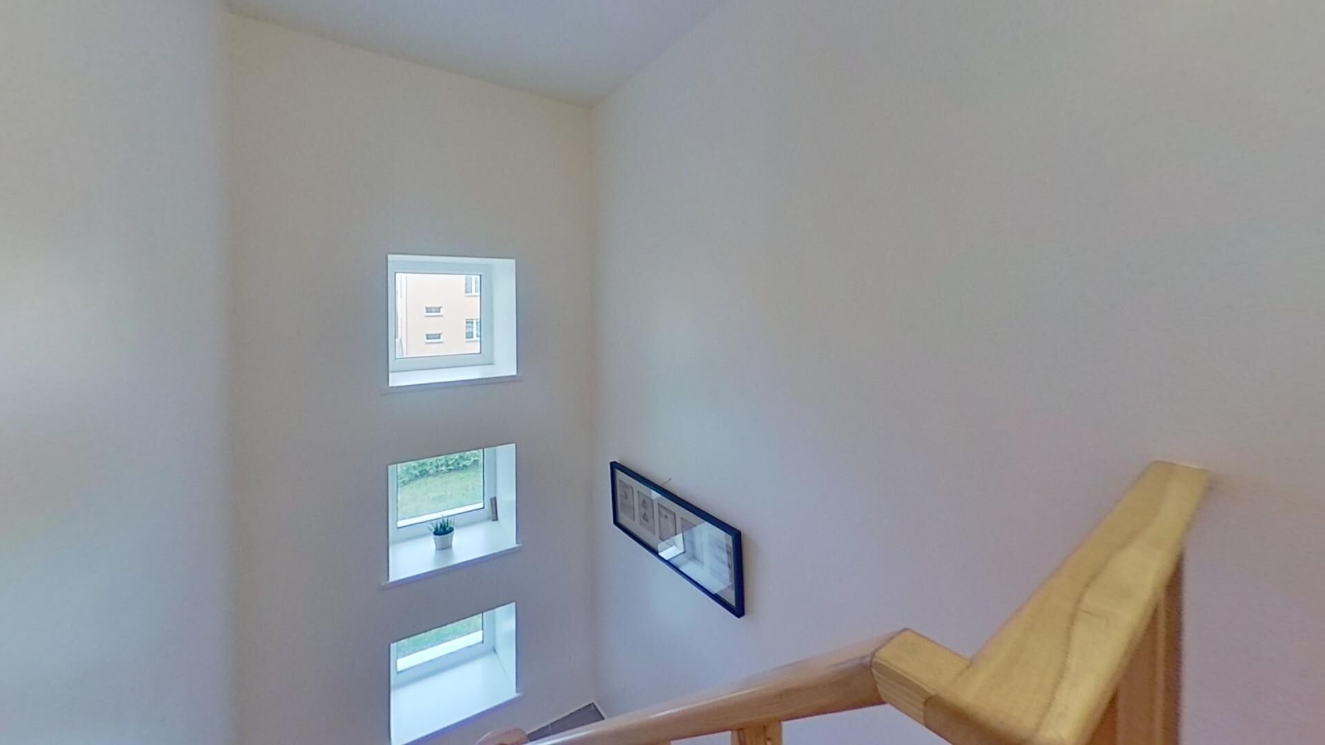 Un escalier avec une rampe en bois mène à un palier. Sur le mur du palier, on trouve trois fenêtres, chacune offrant une vue différente : l’une donne sur un immeuble, une autre sur un jardin, et la troisième est dégagée.