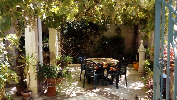 A covered patio area with a table and chairs, surrounded by potted plants and a metal gate.