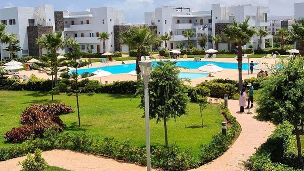 A resort with a central swimming pool, palm trees, and modern white buildings. - Beautiful apartment in La Perla 2 4332 (Saidia)