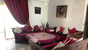 A modern living room with a sectional sofa, a coffee table, and a vase with dried plants. There are two paintings on the walls and a large window with curtains. - Beautiful apartment in La Perla 2 4332 (Saidia)