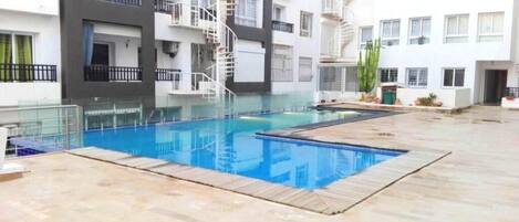 A swimming pool area with a wooden deck, surrounded by a multi-story building with balconies and a spiral staircase.