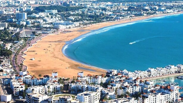 Luchtbeeld van een kuststad met een breed strand, een dichtbebouwd gebied en een heldere blauwe zee.