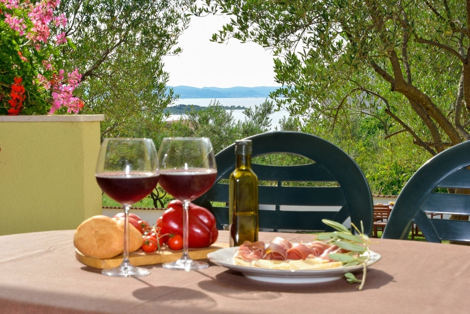 A table with two glasses of red wine, a bottle of olive oil, bread, tomatoes, and a plate of cured meats, set against a backdrop of a scenic lake and trees.