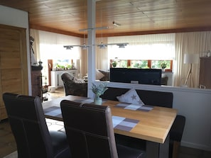Uma área de refeições com mesa de madeira, cadeiras estofadas em preto, e vista para uma cozinha e sala de estar com lareira, plantas em vasos e uma televisão grande.