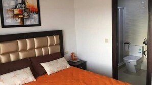 Un dormitorio con cama, una mesita de noche, un cuadro abstracto enmarcado y un baño visible a través de una puerta de cristal.