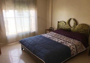 Quarto com cama, uma cadeira e janela com cortinas leves.