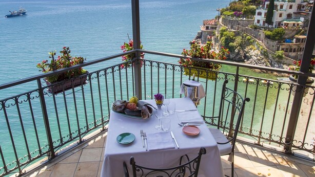 A balcony with a table set for dining, overlooking a coastal view with buildings and the sea. - Hotel Cetus (Cetara)