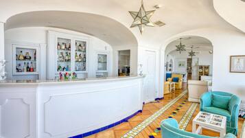 A bar area with a white counter, shelves with bottles, and a patterned floor. There are teal chairs and a white table with a glass.