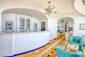 Uma área de bar com balcão branco, prateleiras com garrafas e piso estampado. Há cadeiras cor turquesa e uma mesa branca com um copo.