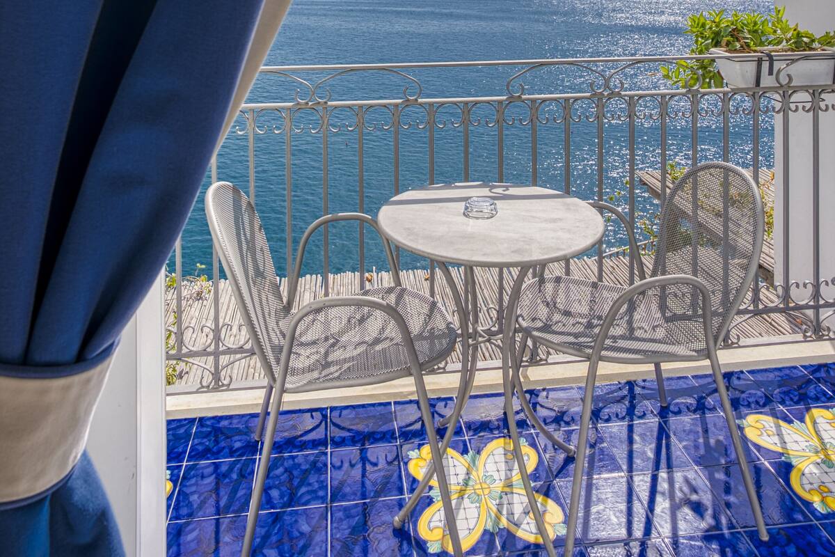 A balcony with a view of the ocean, a small table, two chairs, and a potted plant.