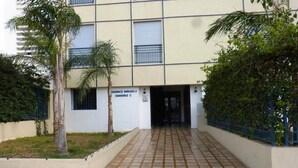 A building entrance with a tiled pathway leading to a door. There are palm trees and a blue fence on the right.