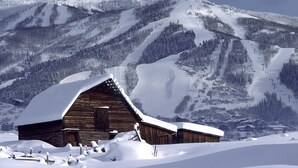 Una cabaña de madera con techo inclinado, rodeada de terreno cubierto de nieve y montañas al fondo.