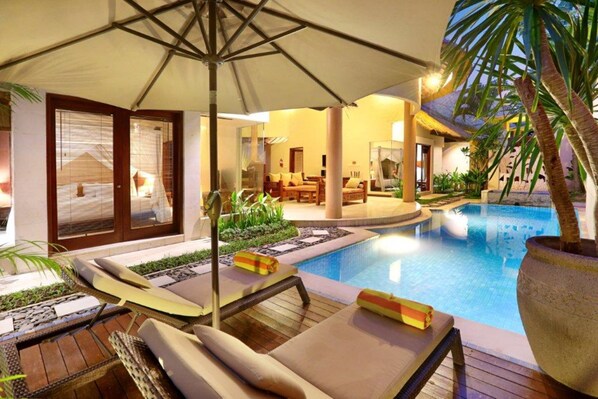 Relax by the pool surrounded by lush greenery and a palm tree, with lounge chairs and a shaded area.