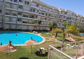 Un hotel con piscina, mobiliario exterior y un edificio de varios pisos con balcones.