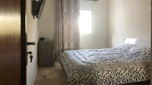 Ein Schlafzimmer mit einem Bett, einem an der Wand befestigten Fernseher, einem Fenster mit Vorhängen und einer Tür mit Schlüssel im Schloss.