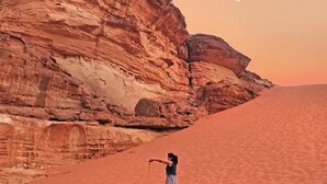 A person standing on a sandy desert with red rock formations and a moon in the sky.