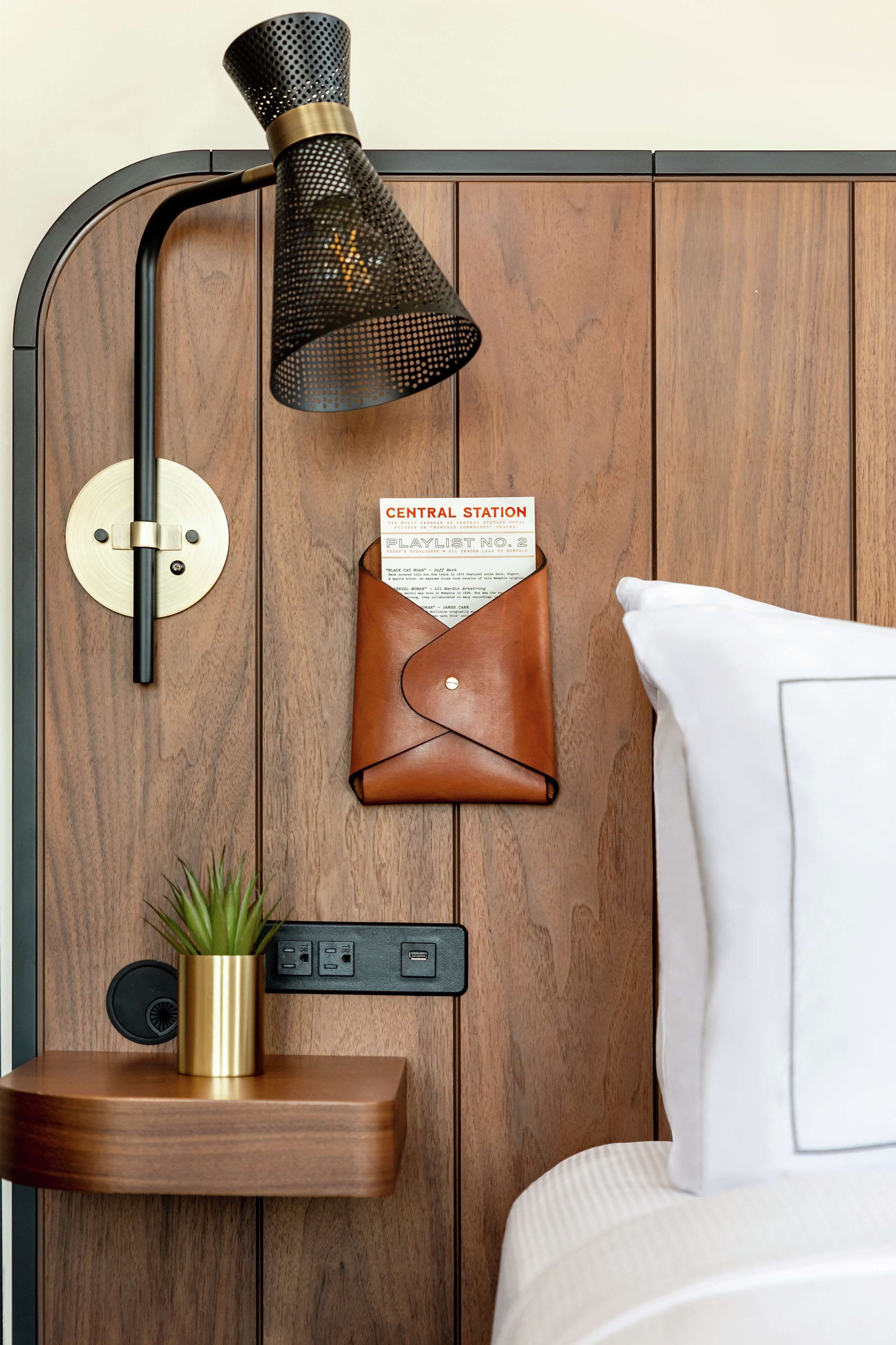 A wooden wall with a black mesh lamp, a leather wallet, a gold-plated plant, and a white pillow.