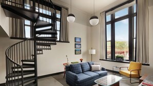 Una sala de estar moderna con un sofá azul, un sillón amarillo, una mesa de centro de vidrio, una escalera espiral negra y ventanas amplias con cortinas.