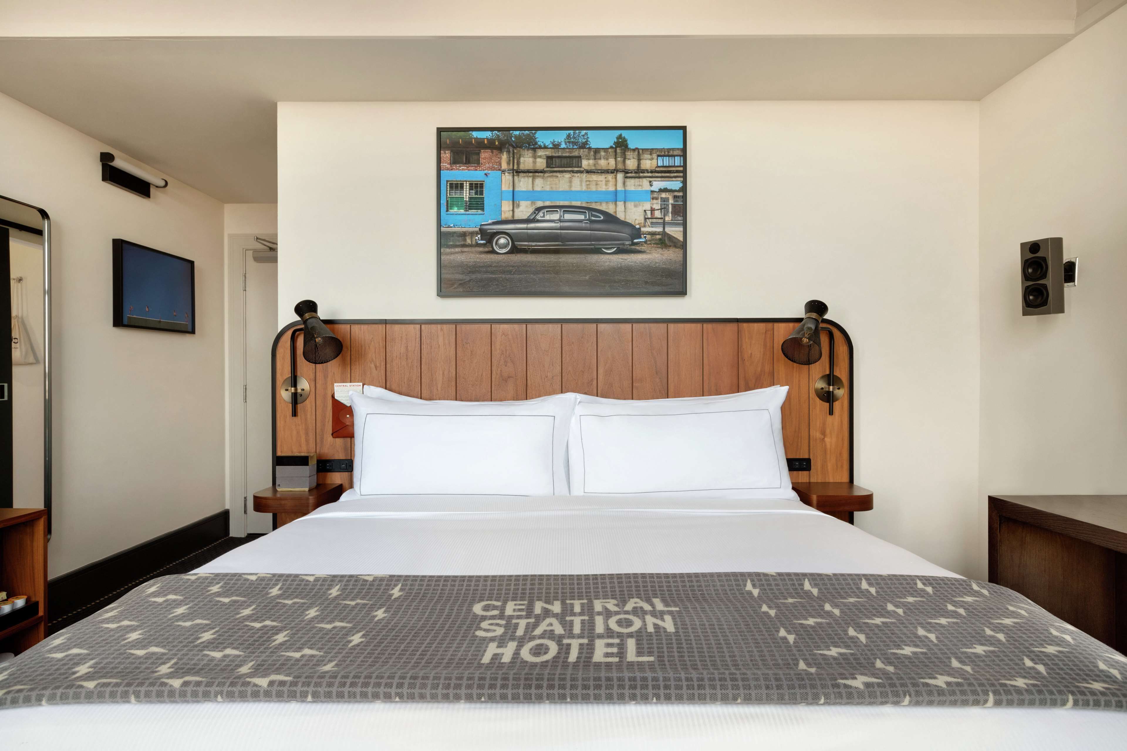 A hotel room with a bed, a headboard with a picture of a car, a wooden nightstand, and a wall-mounted speaker.