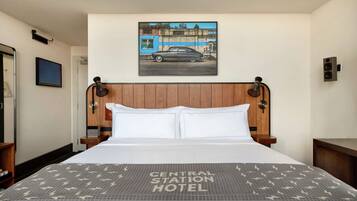 A hotel room with a bed, a headboard with a picture of a car, a wooden nightstand, and a wall-mounted speaker.