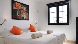 A hotel room with two beds, each with white bedding and orange pillows, a painting on the wall, a window with black frames, and a nightstand with a lamp. - Modern, upstairs apartment with sea view, Wifi, Air Con and U.K. TV (Puerto del Carmen)