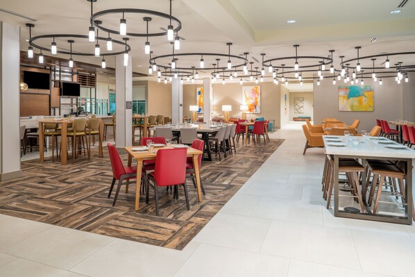 A modern dining area with multiple tables and chairs, overhead lighting, and a herringbone-patterned floor. - Home2 Suites by Hilton Brunswick (Brunswick)