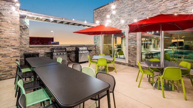 Eine Terrasse im Freien mit einem Grill, grünen und schwarzen Tischen sowie roten Sonnenschirmen.