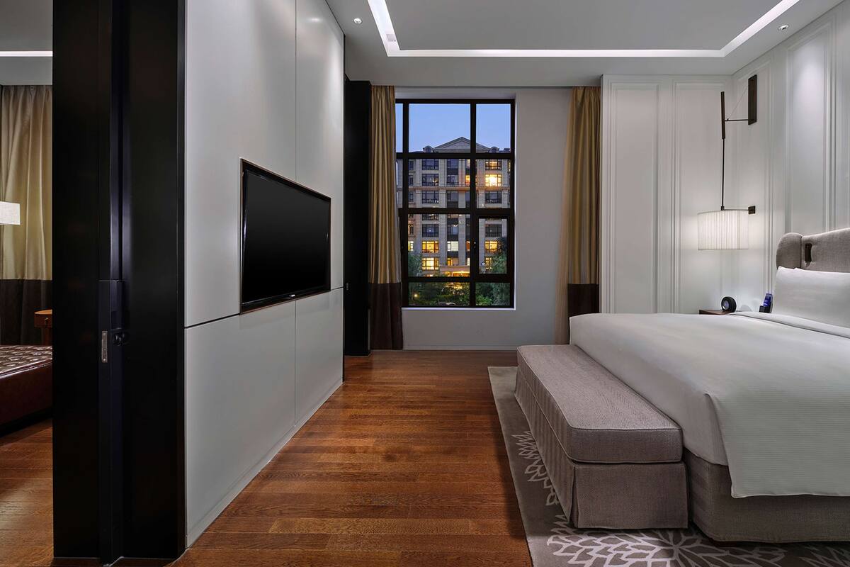 Ein modernes Hotelzimmer mit einem großen Bett, einem flachbildfernseher an der Wand, einer Sitzbank und Blick auf Gebäude durch das Fenster.