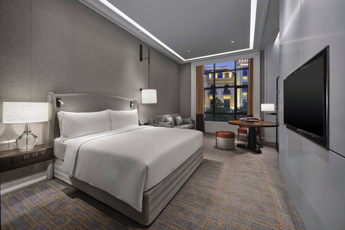 Ein modernes Hotelzimmer mit einem großen Bett, einem Flachbildfernseher, einem Essbereich und Blick auf die Stadtlandschaft durch das Fenster.
