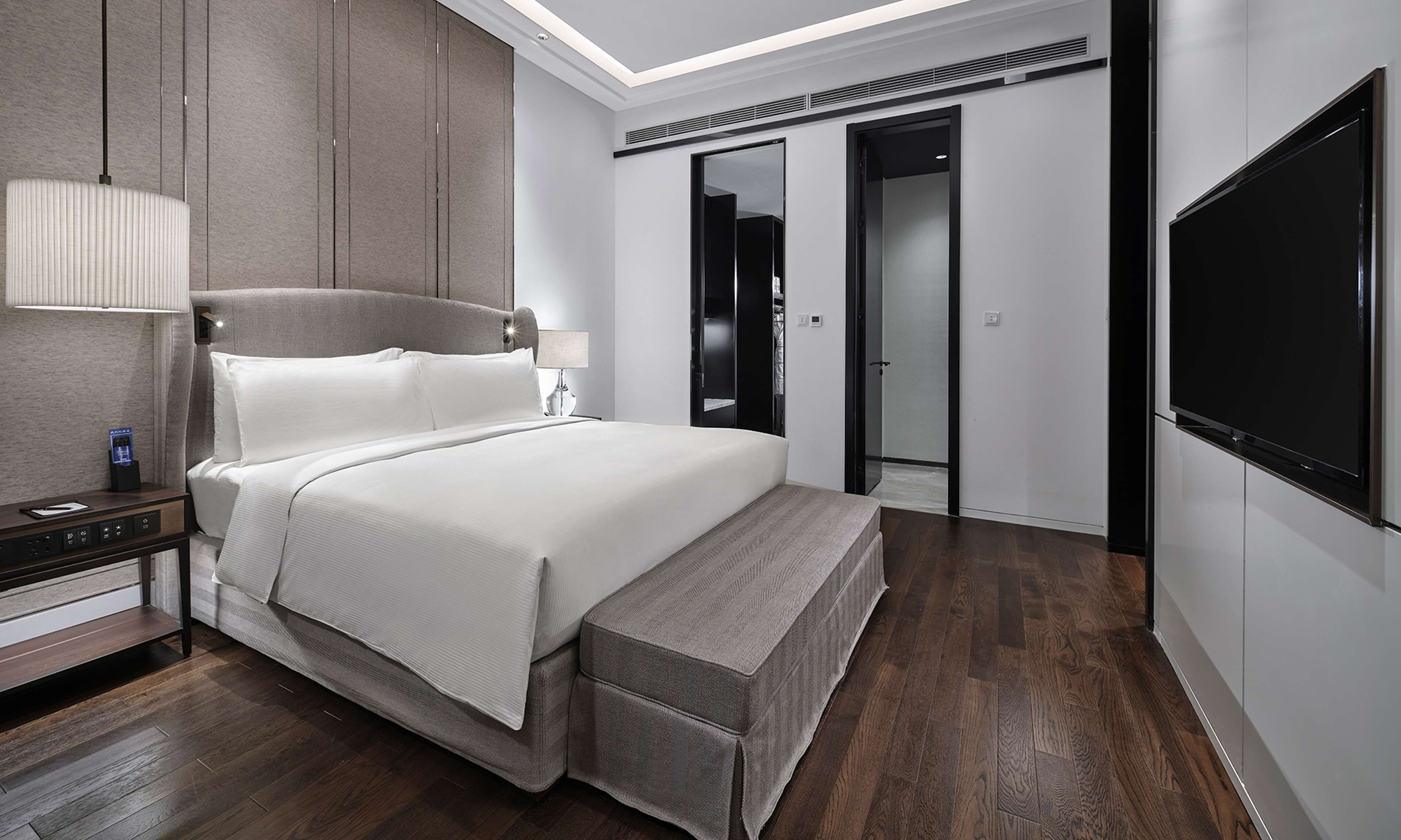 A modern hotel room with a large bed, a bedside table with a lamp, a TV, and a wardrobe.