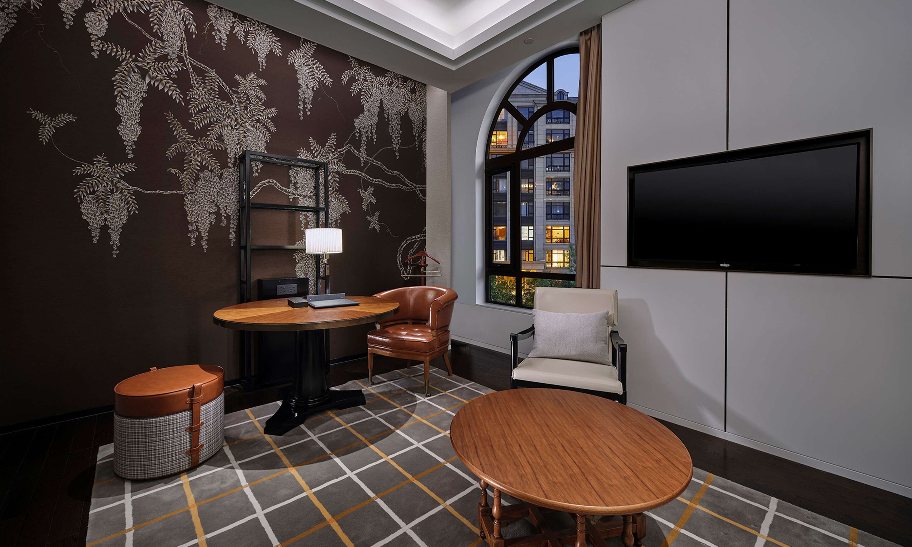 A modern hotel room with a large window, a flat-screen TV, a round wooden table, a leather armchair, and a patterned rug.