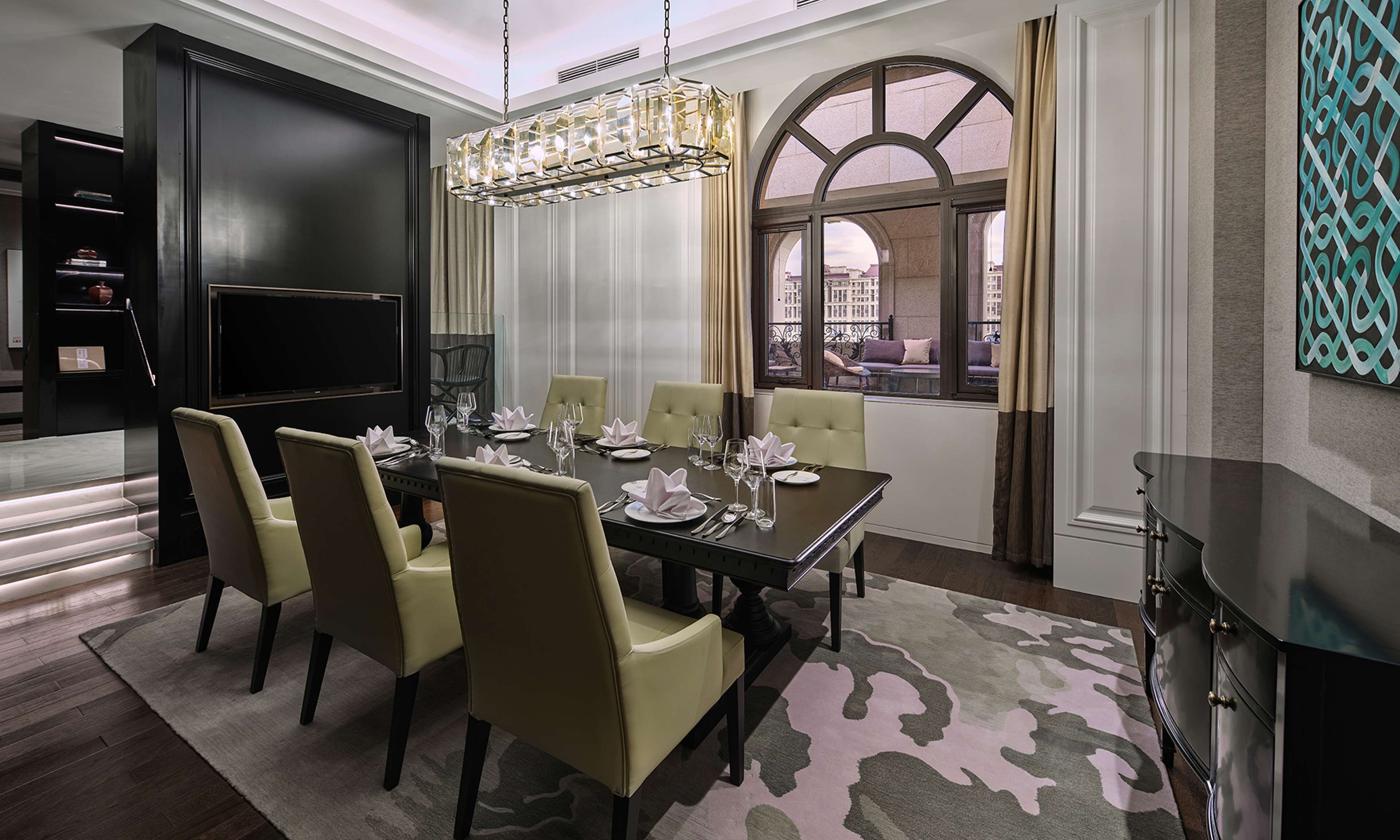 A dining room with a large table set for six, surrounded by six chairs. There is a chandelier above the table, and a large window with curtains on the right side.