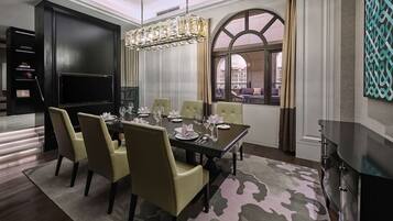 A dining room with a large table set for six, surrounded by six chairs. There is a chandelier above the table, and a large window with curtains on the right side.