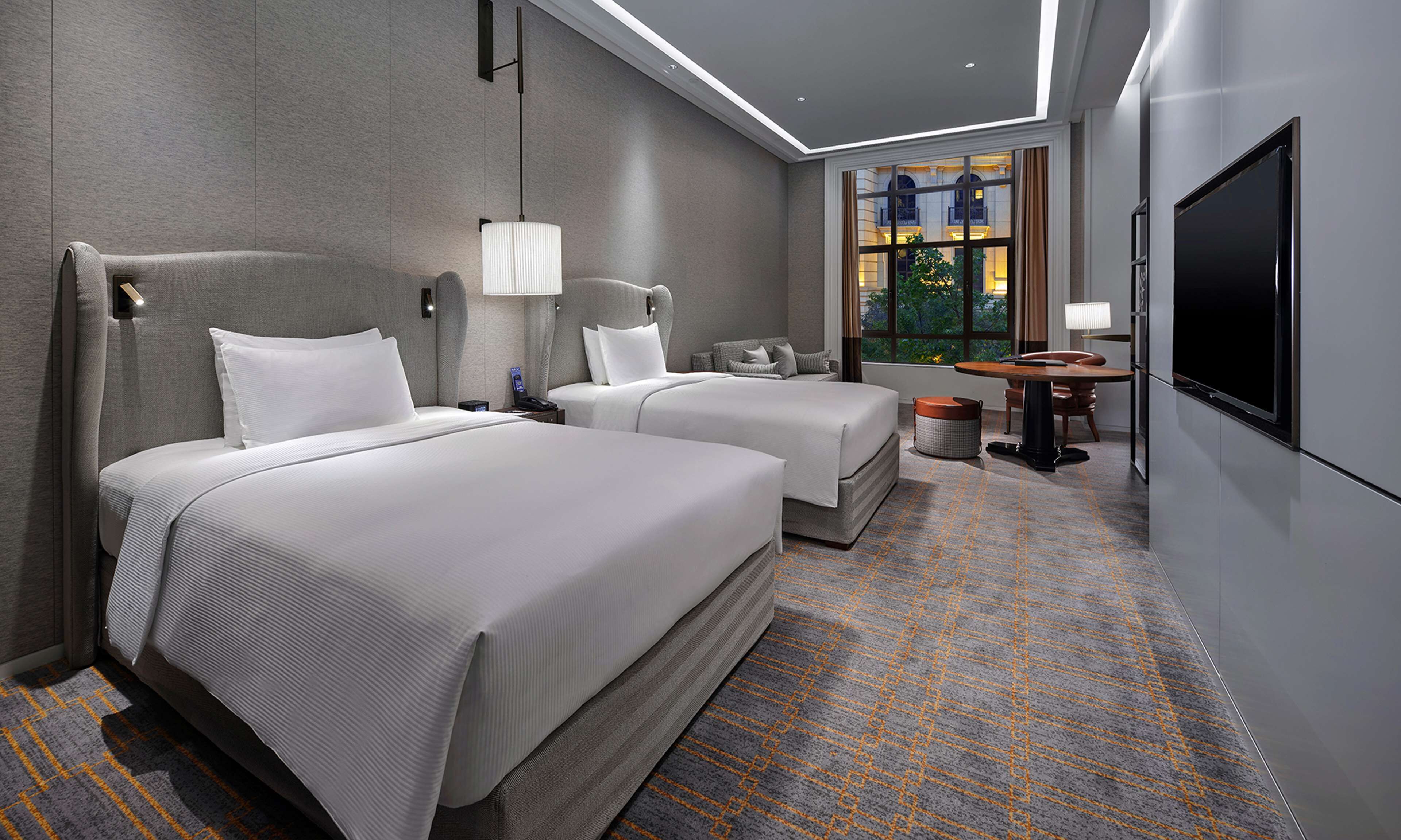 A hotel room with two beds, a TV, a window with a view, and a small table.