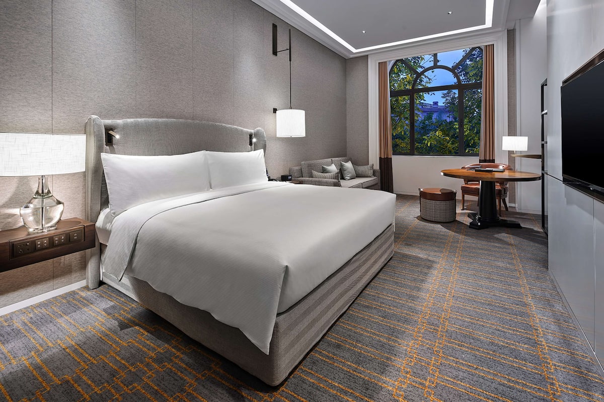 Ein modernes Hotelzimmer mit einem großen Bett, einem Schreibtisch, einem Fernseher und einem Fenster mit Blick auf Bäume.