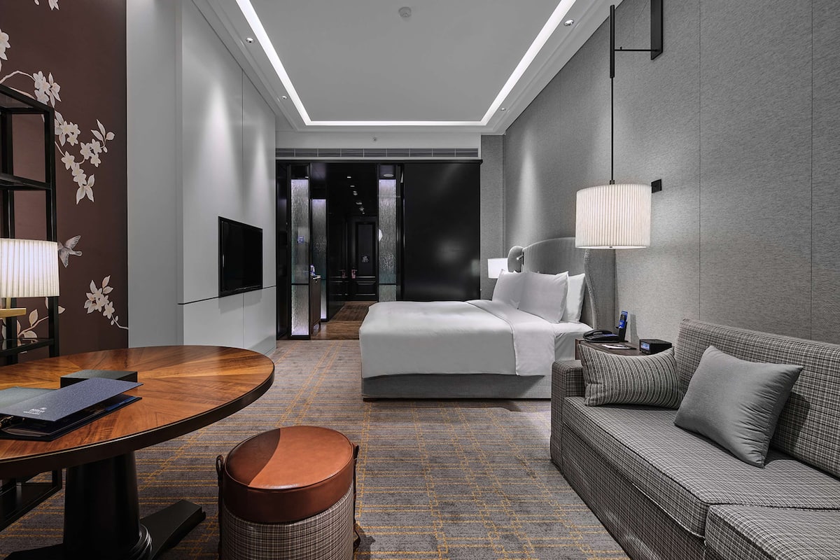 Ein modernes Hotelzimmer mit Bett, Sofa, Schreibtisch mit Lampe, Fernseher und Blick auf den Flur.