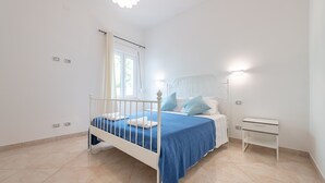 Un dormitorio con cama blanca, ropa de cama azul, una mesita de noche blanca y una ventana con cortinas.