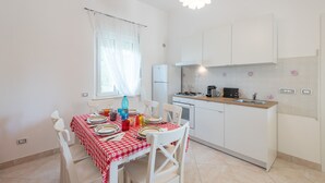 Una cocina compacta con armarios blancos, un pequeño comedor con mantel a cuadros y una ventana con cortinas traslúcidas.