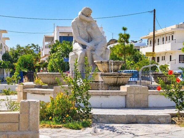 A public square with a classical statue, a fountain, and surrounding greenery.