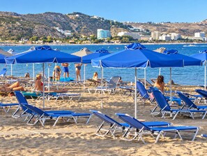 A beach with rows of blue sun loungers and umbrellas, a clear blue sea, and buildings in the background.
